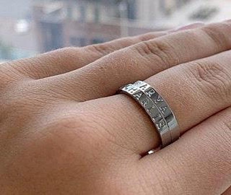 May include: A silver-colored metal ring featuring two stacked bands. The top band is engraved with the name ARYA and the bottom band is engraved with the name CHARLIE. The ring has a smooth, polished finish and a simple, clean design.
