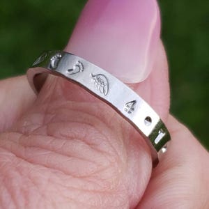 May include: A silver-toned metal ring featuring engraved symbols along its outer band. The visible engravings include small footprints, a stylized wing, the number four, and a circular dot. The band has a smooth, polished finish throughout.