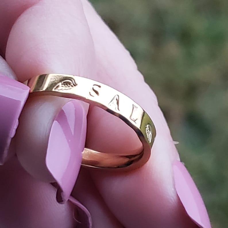 Puede incluir: Un anillo de oro con la palabra "SAL" grabada. El anillo est&aacute; siendo sostenido por una mano con esmalte de u&ntilde;as rosa.