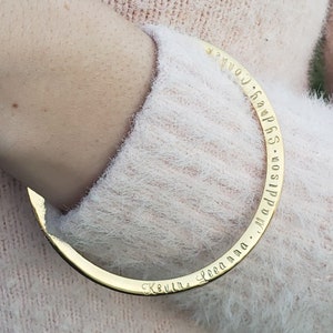 May include: A gold-colored bangle bracelet with engraved names. The bracelet is worn on a wrist covered by a light pink, fuzzy sweater. The names engraved on the bracelet include Kevin, Leeanna, Maddison, Sydney, and Connor.