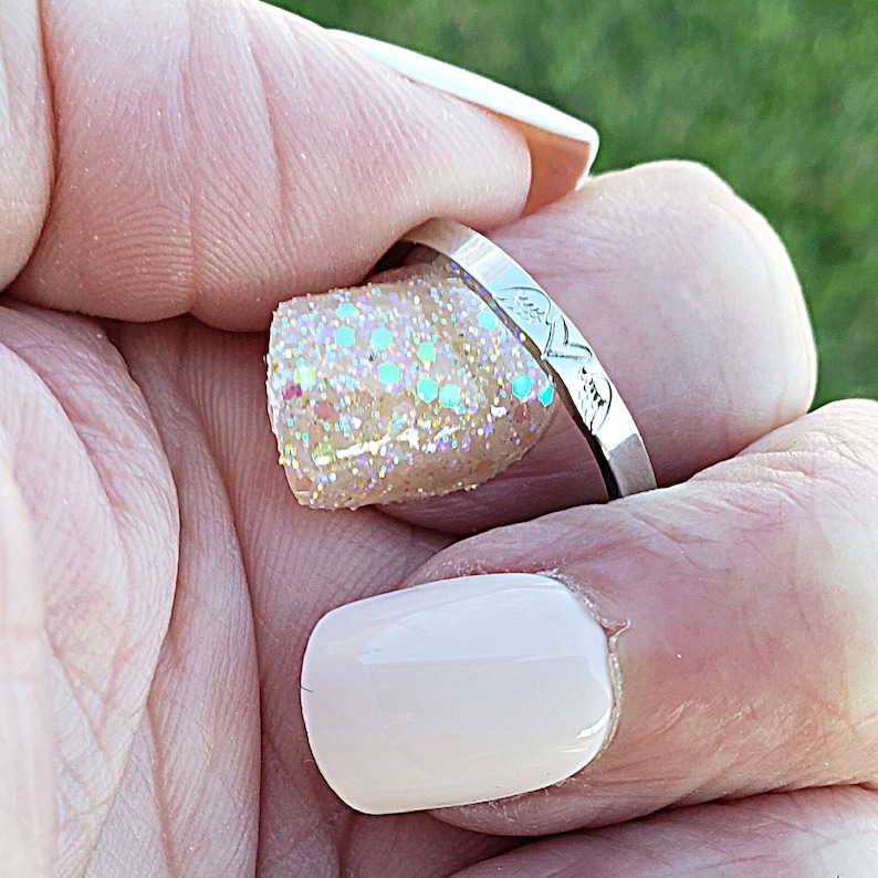 May include: A silver ring with a delicate design of two hearts intertwined. The ring is worn on a finger with a light pink nail polish and a glittery nail polish.