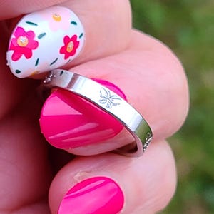 May include: A silver ring with a butterfly design engraved on the band. The ring is on a finger with pink nail polish.