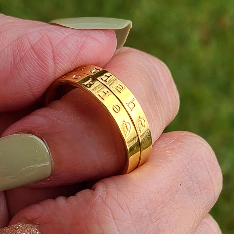 May include: Two gold-toned metal rings featuring engraved lettering and small leaf-like symbols. The bands have a smooth, polished finish and are stacked together, displaying the text "h e l l o" clearly etched into the surface of the metal bands.