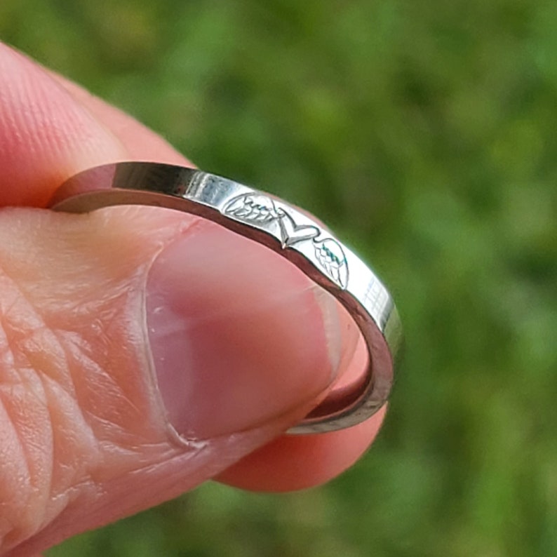 May include: A silver ring with a heart and wings design. The wings are detailed with a small line of green enamel.