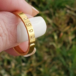 May include: A gold-toned metal ring with a smooth, flat surface featuring the engraved numbers 1965. The band has a consistent width and a polished finish, showcasing the stamped digits clearly along the outer circumference of the circular jewelry piece.
