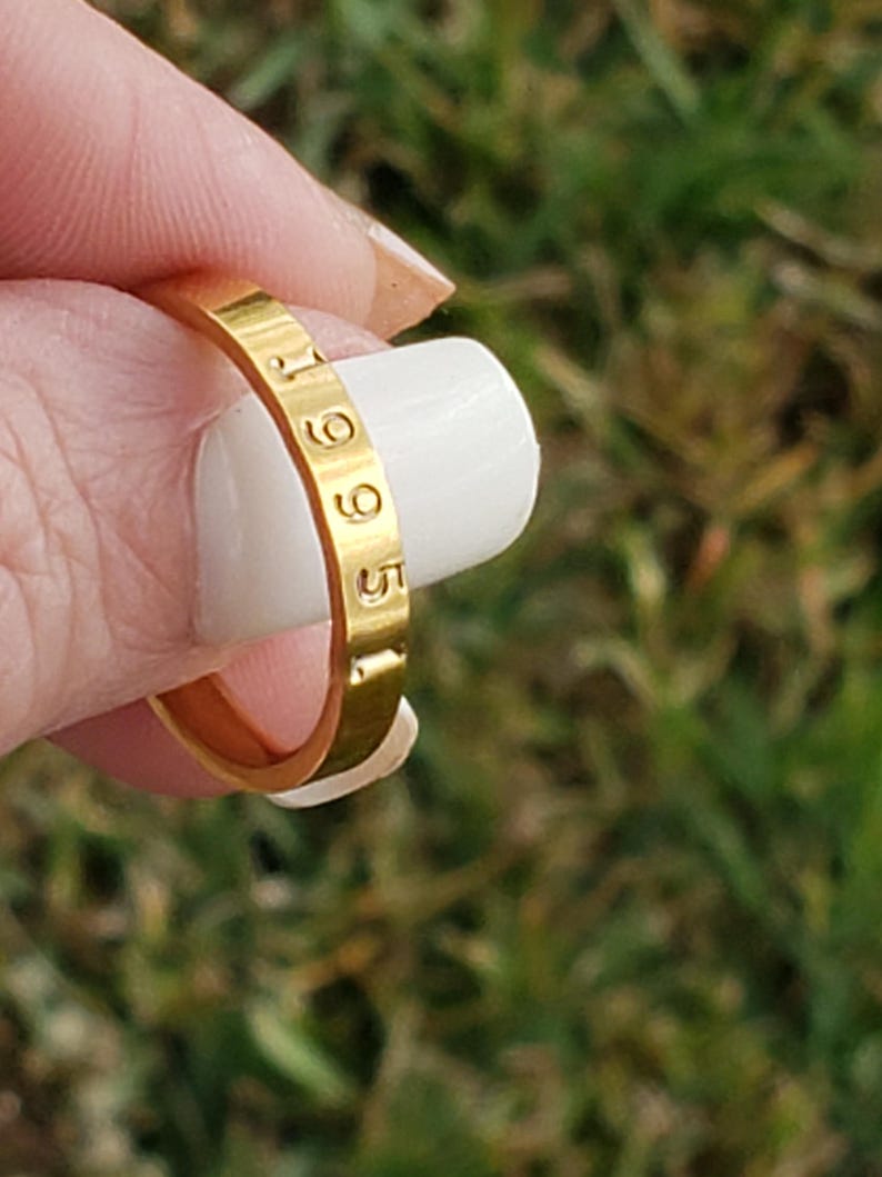 May include: A gold-toned metal ring with a smooth, flat surface featuring the engraved numbers 1965. The band has a consistent width and a polished finish, showcasing the stamped digits clearly along the outer circumference of the circular jewelry piece.