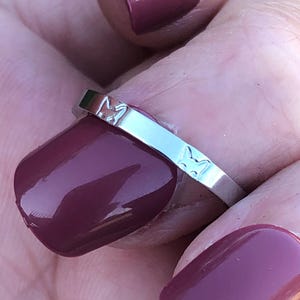 May include: A polished silver-tone metal ring featuring a repeating embossed pattern of small cat faces along the outer band. The ring has a smooth, reflective surface and a slim, uniform width, showcasing a simple design with distinct feline silhouettes.