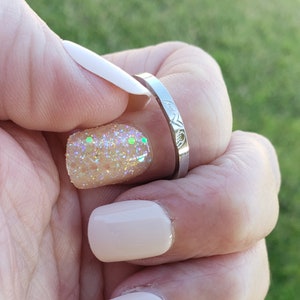 May include: A silver ring with a delicate design on the band. The ring is on a finger with a light pink nail polish and a glittery nail polish.