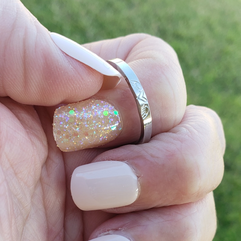 May include: A silver ring with a delicate design on the band. The ring is on a finger with a light pink nail polish and a glittery nail polish.