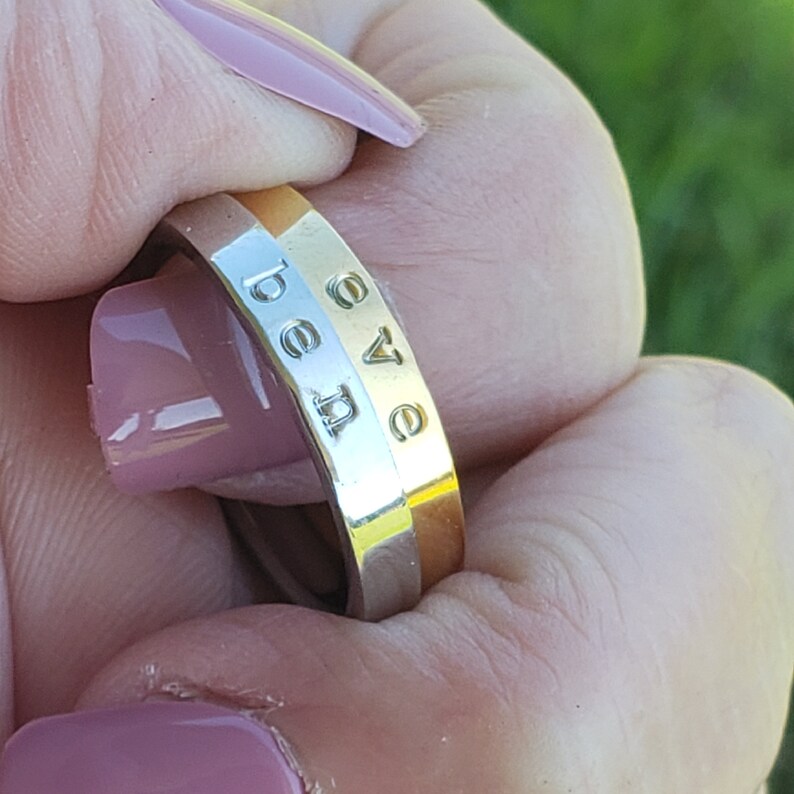 May include: A gold and silver ring with the word "be love" engraved on the band. The ring is held up against a green background.