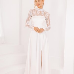 May include: A white maxi dress with a lace long-sleeve top and a high neckline. The dress has a fitted bodice and a flowing skirt with a slit in the front.