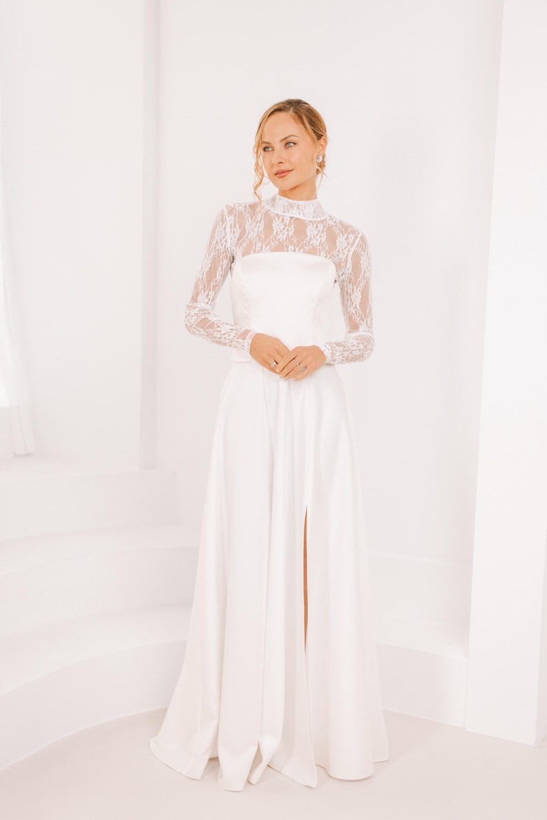 May include: A white maxi dress with a lace long-sleeve top and a high neckline. The dress has a fitted bodice and a flowing skirt with a slit in the front.