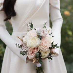 Könnte beinhalten: Ein Hochzeitsstrauß mit weißen Rosen, rosafarbenen Nelken und hellrosa Rosen, mit grünem Laub. Der Strauß wird vor einem weißen Kleid mit langen Ärmeln gehalten. Die Blumen sind mit einem weißen Band gebunden.
