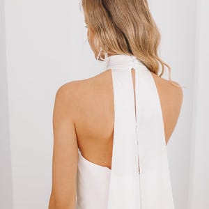 May include: A white wedding dress with a high, halter-style neckline and a flowing back detail. The dress has a keyhole back and a long, elegant drape. The model's blonde hair is styled in loose waves, and she wears delicate earrings.