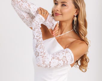 May include: A pair of white lace arm sleeves featuring a detailed floral pattern. The sleeves extend from the wrist to the upper arm, crafted from a delicate, semi-sheer fabric with scalloped edges, worn over a plain white sleeveless dress.