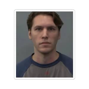 May include: A sticker featuring a portrait of a person with light skin and brown hair. The person is wearing a gray and blue baseball style shirt. The background is a solid gray color.