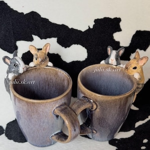 Może przedstawiać: Dwa ceramiczne kubki z nakrapianą brązowo-szarą glazurą, każdy z dwoma małymi rzeźbionymi królikami wyglądającymi znad krawędzi. Napis julii.sk.art jest widoczny na boku każdego kubka. Króliki są pomalowane w odcieniach szarości, bieli i beżu.