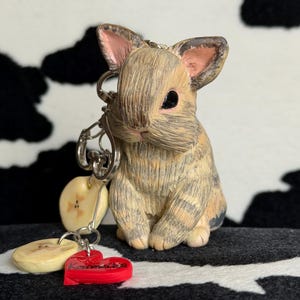 Op de afbeelding: Een sleutelhanger met een gebeeldhouwd beige en grijs konijn met donkere ogen en roze gevoerde oren. Aan de zilveren metalen ring hangen twee kleine, ronde, crèmekleurige bedels en een rood hartvormig bedeltje met de naam Kamila erop gedrukt.