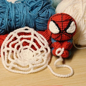 Op de afbeelding: Een gehaakt Spider-Man-figuur van rood en blauw garen met grote witte ogen en zwarte webdetails, vergezeld van een wit gehaakt spinnenweb-accessoire. Het figuur is geplaatst voor verschillende kleurrijke bollen garen.