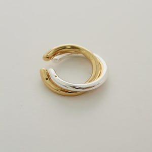 May include: A two-tone ear cuff featuring a gold and silver intertwined design. The ear cuff has a smooth, polished finish and a minimalist aesthetic. The jewelry is designed to be worn on the ear without piercing.