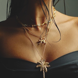 May include: A collection of gold necklaces with various pendants, including a palm tree and a small animal. The necklaces are layered and worn against a dark background, creating a contrast that highlights the jewelry's details.