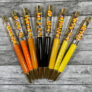 May include: Set of eight gold-capped pens with clear barrels filled with candy corn. The pens have different colored glitter accents.