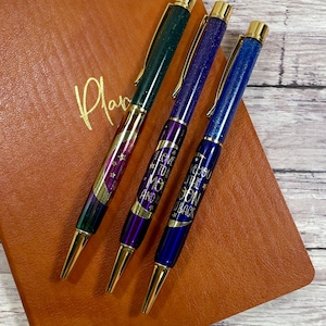 May include: Three pens with gold accents and glitter embedded in the ink. The pens have different designs, including a crescent moon and stars, and the text "I love you to the moon and back".