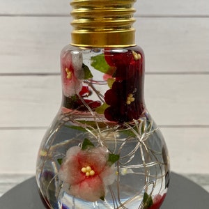 May include: A clear glass light bulb with a gold metal base filled with water, red and pink flowers, and silver wire lights.