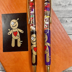 May include: Two gold-toned pens with clear barrels filled with colorful beads and glitter. The pens have a voodoo doll design with a red heart and black eyes. One pen has a black base and the other has a purple base.