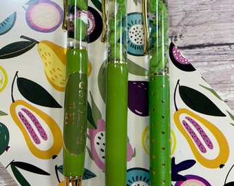 May include: Three green pens with gold accents and clear barrels filled with green glitter. The pens have different designs, one with a gold heart pattern, one with a gold polka dot pattern, and one with a gold inscription that reads "Be Kind".