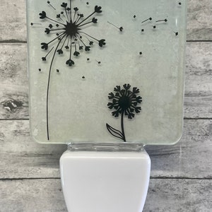 May include: A white night light with a glass panel featuring a black silhouette of two dandelions. The larger dandelion has seeds blowing in the wind.
