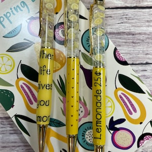 May include: Three yellow pens with gold accents and clear bodies filled with lemon slices. The pens have different sayings on them: "When life gives you lemons", "Lemonade 25¢", and a pen with yellow hearts.