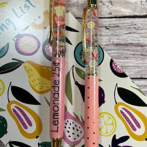 May include: Two pink pens with gold accents and clear barrels filled with colorful fruit slices. One pen has the text "Lemonade 25¢" printed on the side.