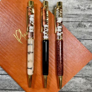 May include: Three gold-capped pens with clear barrels filled with glitter and white beads. The pens have different designs: one with black ink and a coffee-themed design, one with gold ink and a glitter design, and one with black ink and a glitter design.
