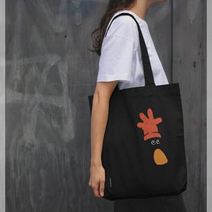 Puede incluir: Bolso tote negro con un diseño de dibujos animados en naranja y blanco. El gráfico presenta una forma de mano, dos ojos y una lágrima. El bolso se lleva al hombro.