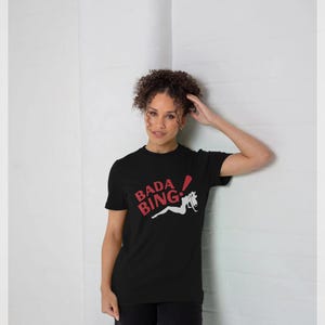 Può includere: T-shirt nero con le parole "BADA BING" in rosso e bianco. La maglietta presenta una grafica di una donna in posizione reclinata. La maglietta è realizzata in un materiale morbido ed è adatta per l'abbigliamento casual.