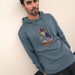 May include: A slate blue hooded sweatshirt featuring a graphic print of the anime character Yugi Muto from Yu-Gi-Oh! on the chest. The hoodie includes a drawstring hood, long sleeves, and a front kangaroo pocket, with the Yu-Gi-Oh! logo printed below.