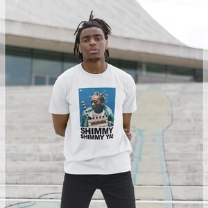 Puede incluir: Camiseta blanca con un gráfico de una persona sosteniendo una claqueta de cine. El gráfico incluye el texto "Shimmy Shimmy Ya!" en negrita, en negro. La persona lleva pantalones negros.