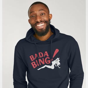 Può includere: Felpa con cappuccio blu navy con la scritta "BADA BING" in rosso e una silhouette bianca di una donna. La felpa ha un cappuccio con coulisse e una tasca a marsupio. La persona nella foto sta sorridendo.