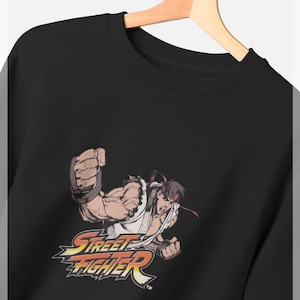 Op de afbeelding: Zwarte sweatshirt met een afbeelding van een Street Fighter-personage in een vechthouding. Het personage draagt een witte gi en een rode hoofdband. Het Street Fighter-logo staat eronder in geel en rood.