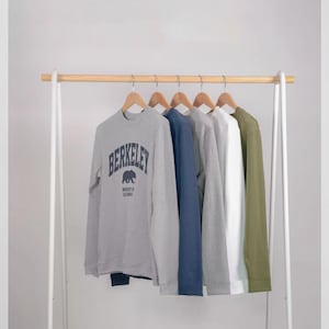 Peut inclure: Un portant blanc avec une barre en bois présente six sweatshirts de couleurs variées. On distingue un sweat-shirt gris avec le texte "BERKELEY", bleu marine, gris clair, blanc et vert olive. Chaque vêtement est suspendu à un cintre en bois.