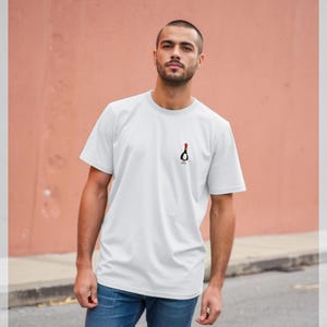 Puede incluir: Camiseta blanca con un pequeño gráfico negro y rojo en el pecho. El gráfico es una botella estilizada con una tapa roja. La persona lleva vaqueros azules. La camiseta tiene mangas cortas.