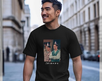 May include: Black t-shirt featuring a rectangular image of two men wearing party hats. The image is from the TV show "Friends", with the show's name printed in white below the image. The t-shirt is worn by a person.