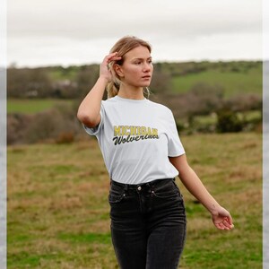 May include: White t-shirt with the text "MICHIGAN Wolverines" in gold and navy blue. The shirt is short-sleeved and has a crew neck. The person is wearing black jeans.