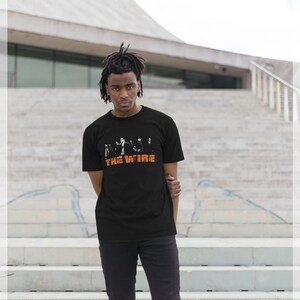 May include: Black t-shirt with an orange "THE WIRE" logo and a graphic of figures. The t-shirt is worn by a person standing in front of a concrete staircase.