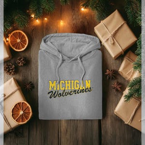 Könnte beinhalten: Ein grauer Kapuzenpullover mit dem Text "MICHIGAN Wolverines" in Gelb und Marineblau. Der Pullover liegt auf einer Holzoberfläche, umgeben von verpackten Geschenken, Tannenzapfen und getrockneten Orangenscheiben.