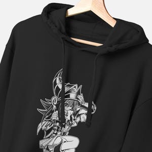 Pode incluir: Uma camisola preta com capuz apresentando uma estampa gráfica branca de uma personagem estilo anime usando um chapéu pontudo e segurando um cajado. O capuz inclui um cordão preto correspondente e está pendurado num cabide de madeira.
