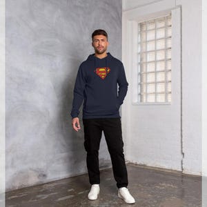 Puede incluir: Sudadera con capucha azul marino con el logotipo de Superman en rojo y amarillo. El logotipo est&aacute; centrado en el pecho. La sudadera tiene un bolsillo delantero y una capucha con cord&oacute;n. La persona lleva pantalones negros y zapatillas blancas.