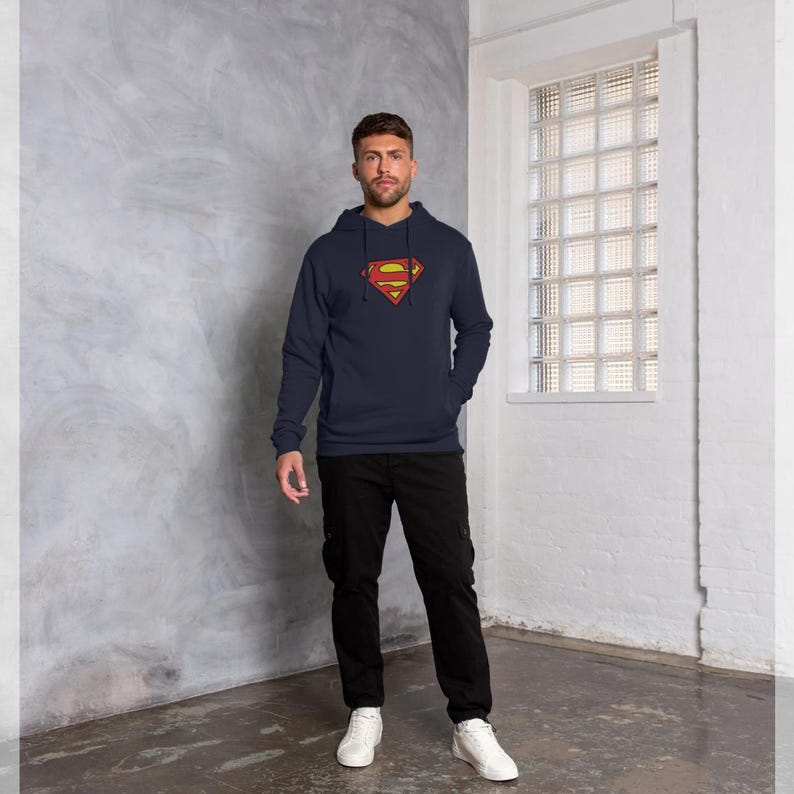 Puede incluir: Sudadera con capucha azul marino con el logotipo de Superman en rojo y amarillo. El logotipo est&aacute; centrado en el pecho. La sudadera tiene un bolsillo delantero y una capucha con cord&oacute;n. La persona lleva pantalones negros y zapatillas blancas.