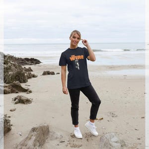 Puede incluir: Una camiseta azul marino con la palabra "Wire" en letras naranjas y un gráfico en blanco y negro. La persona lleva pantalones negros y zapatillas blancas. El fondo es una playa con rocas y el océano.